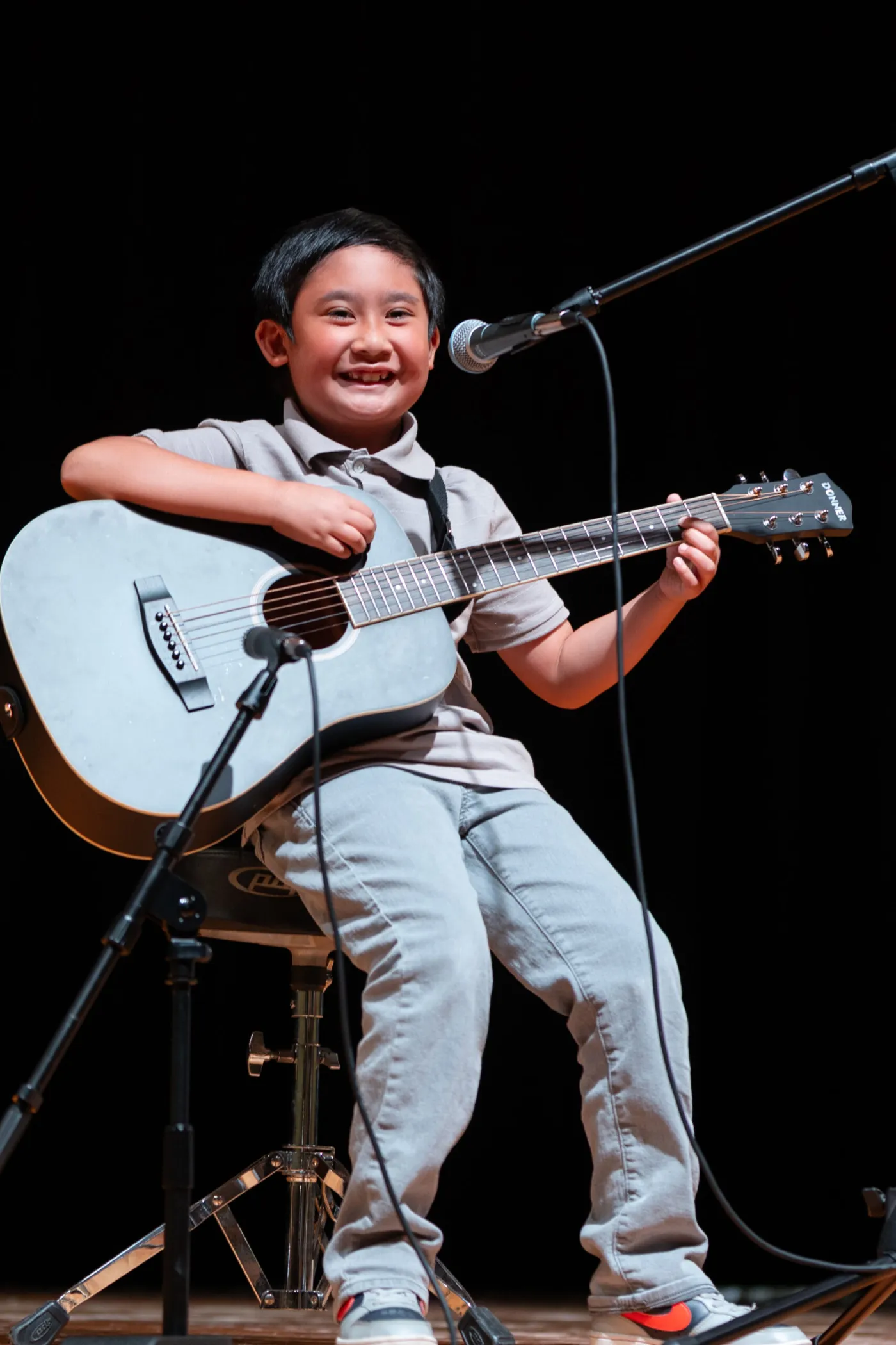 Young guitar student smiling while playing acoustic guitar on stage at a LyriCo recital
